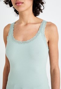 Canotta a coste verde menta chiaro con pizzo lungo il collo e le spalle. Il tessuto appare morbido con un design aderente.