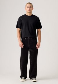 Homme debout portant un t-shirt noir à manches courtes, un pantalon large en velours noir, des baskets noir et blanc, un bracelet en argent et une bague.