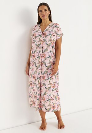 Femme debout pieds nus contre un mur blanc, portant une robe midi boutonnée à manches courtes rose à fleurs.