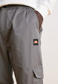 Grå cargobyxor med elastisk midja, sidofickor och en svart lapp med en design i gult, rött och vitt. Mjuk tygtextur.