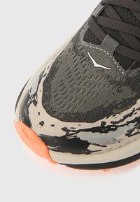 Γκρι αθλητικό παπούτσι με υφασμάτινη επιφάνεια από πλέγμα με υφή και πολύχρωμα σχέδια με πιτσιλιές μπογιάς, με πορτοκαλί σόλα και λευκές λεπτομέρειες με λογότυπο.