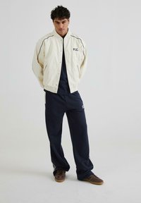 Off-white bomberjakke med marineblå kant og broderet "P.S.C." logo, parret med marineblå sweatpants og brune sneakers. Glat tekstur.