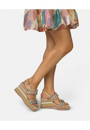 Jambes portant une jupe florale multicolore et des sandales compensées avec des brides perlées et une plateforme tissée, sur fond uni.