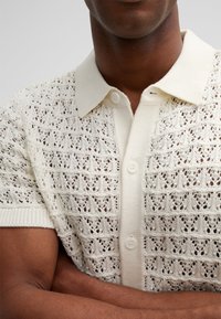 Strickpolohemd mit kurzen Ärmeln in Créme mit einem Spitzenmuster, ausgestattet mit einem weichen Kragen und einer Knopfleiste mit drei Knöpfen. Mit einem Strukturmuster aus Durchbrüchen versehen.