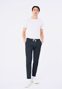 T-shirt blanche à manches courtes associée à un pantalon chino ajusté bleu marine avec taille à cordon, complétée par des baskets blanches. Fond neutre.