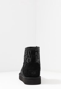 Botas negras hasta el tobillo hechas de ante, con un textil estampado que presenta el nombre de la marca. Tienen una suela de goma plana y un tirador en la parte trasera.