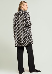 Zwart- en crèmekleurig golvend patroon lang jack van gestructureerde stof, gecombineerd met zwarte broek en platte schoenen. Eenvoudig kraagontwerp.