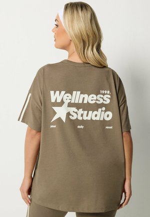 Blonde Frau trägt übergroßes braunes T-Shirt mit weißem Text „Wellness Studio 1998“ und Stern-Grafik auf dem Rücken, stehend vor einfarbigem Hintergrund.