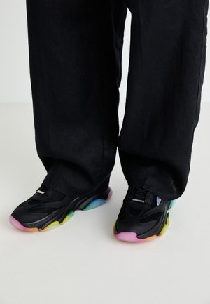 Baskets noires avec une tige en maille, semelles en caoutchouc colorées dans des teintes de rose, jaune et bleu, avec un design épuré et arrondi.