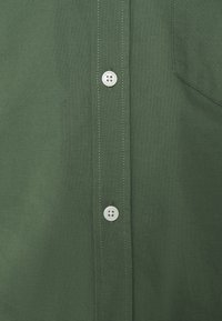 Groene shirt gemaakt van stof met een gladde textuur, voorzien van een knoopsluiting en een borstzak, details met contrasterende witte stiksel.