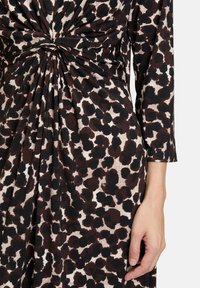 Gros plan sur une robe à imprimé léopard avec un détail de nœud froncé à la taille, montrant une longue manche et la main droite reposant sur le côté.