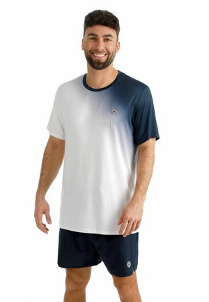 Lachende man die een marineblauw en wit gradient T-shirt met korte mouwen en marineblauwe korte broek draagt, staand tegen een witte achtergrond.