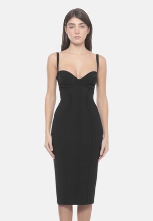 Donna in piedi davanti a uno sfondo semplice, indossando un vestito nero aderente con spalline, busto strutturato e orlo all'altezza del ginocchio.