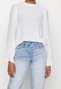 Blouse blanche texturée à manches longues avec un col à volants, associée à un jean en denim bleu clair taille haute à coupe droite.