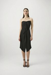 Crna haljina bez naramenica s naborima na prednjem dijelu; duljine do koljena s šiljastim rubom; glatka tkanina. Nošena s crnim sandalama s remenčićem oko gležnja.