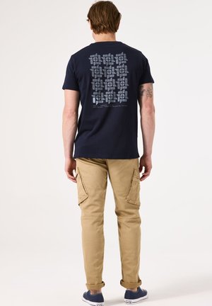 Marine t-shirt met een grafisch patroon op de rug, gecombineerd met beige cargobroeken met zijzakken en blauwe sneakers.