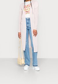 Light pink long cardigan over a white floral t-shirt, paired with light blue flared jeans and white platform sneakers. Beige crossbody bag.