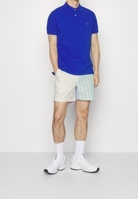 Blå polotröja med korta ärmar och en grön logotyp, tillsammans med randiga shorts i pastellfärger. Vita sneakers med grå detaljer.