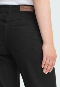 Černé džínsy z denimu s hnědou koženou nášivkou na pasu, jednou zadní kapsou a hladkým povrchem.