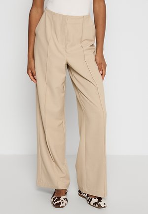 Pantalon beige taille haute à jambes larges porté avec des chaussures plates à imprimé vache noir et blanc, présenté de la taille aux pieds sur un fond uni.