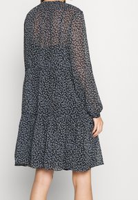 Mujer que lleva un vestido negro hasta la rodilla con mangas largas translúcidas y un pequeño patrón floral azul claro, vista desde la espalda.