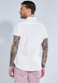 Weißes Kurzarmhemd mit Button-Down-Kragen, aus strukturiertem Stoff und grauen Akzenten an den Ärmeln, kombiniert mit hellrosa Hosen.