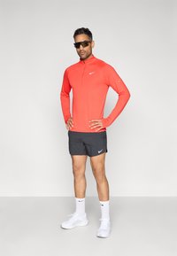 Κόκκινο μπλουζάκι μακρυμάνικο της Nike με φερμουάρ μέχρι τη μέση, μαύρο αθλητικό σορτς, λευκές κάλτσες και παπούτσια. Ανάκλαστικά στοιχεία και λογότυπο στα ρούχα.