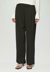 Schwarze maßgeschneiderte Hosen mit hoher Taille, gefälteltem Vorderteil und weitem Bein. Der Stoff hat eine glatte Textur. Kombiniert mit schwarzen Riemchenheels.