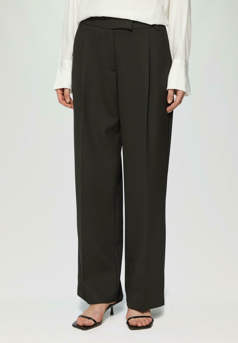 Schwarze maßgeschneiderte Hosen mit hoher Taille, gefälteltem Vorderteil und weitem Bein. Der Stoff hat eine glatte Textur. Kombiniert mit schwarzen Riemchenheels.