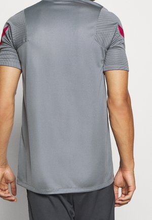 Mann trägt ein kurzärmeliges graues Sportshirt mit strukturierten Schultern und roten Akzenten an den Ärmeln, von hinten gezeigt.