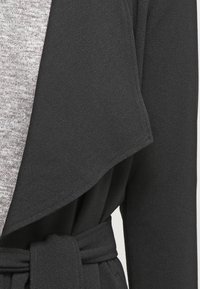 Musta kietaiscoat, jossa on vyöllinen vyötärö, pehmeä rakenne ja leveä kaulus, joka peittää alla olevan harmaan neuleen.