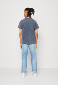 Camiseta de algodón gris con un corte relajado, mangas enrolladas, combinada con vaqueros de denim de lavado claro; el modelo lleva zapatos blanco tipo slip-on.