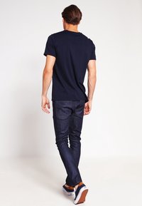 Marinblå bomulls-T-shirt med kort ärm, kombinerad med mörkblå denimjeans. Modellen ses från baksidan, iklädd avslappnade skor med vita sulor.