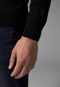 Sort strikket sweater med ribbede manchetter, der viser et nærbillede af en hånd, der hviler på mørk denimstof. Teksturen ser glat ud.