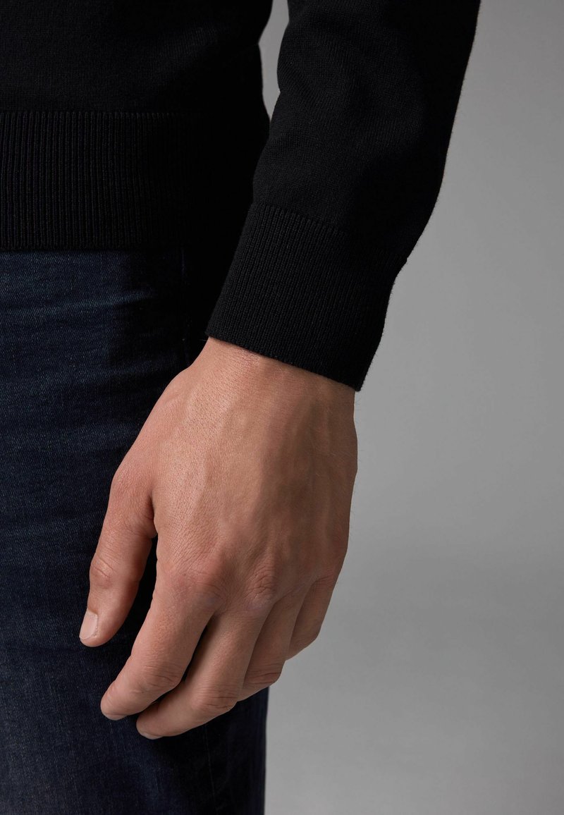 Sort strikket sweater med ribbede manchetter, der viser et nærbillede af en hånd, der hviler på mørk denimstof. Teksturen ser glat ud.