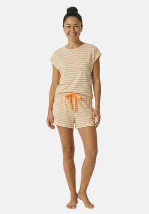 Femme debout pieds nus portant un haut à manches courtes rayé beige et orange et un short assorti avec un cordon orange, souriant à la caméra.