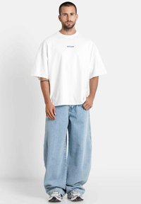 Witte oversized t-shirt met blauwe logo, gecombineerd met lichtblauwe wijde spijkerbroek en grijze sneakers. Gladde stof en losse pasvorm.