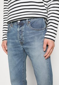 Vaqueros de mezclilla azul claro con acabado desteñido, cinco bolsillos y herrajes de cobre. Combinados con una camiseta de manga larga a rayas en blanco y negro.