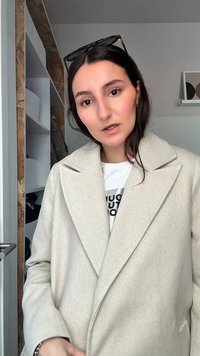 Žena s tmavými vlasy, která má na sobě béžový kabát a brýle na hlavě, stojí uvnitř poblíž otevřené skříně s policemi a oblečením.