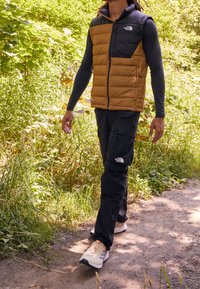 Vadderad väst med bruna och svarta sektioner, grå långärmad tröja, svarta cargobyxor och löparskor på en grusväg omgiven av grönska.