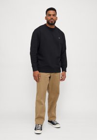 Svart sweatshirt med rund hals och ribbade ärmar, kombinerad med beige chinos och svarta sneakers med vita sulor. Enkelt, avslappnat outfit.