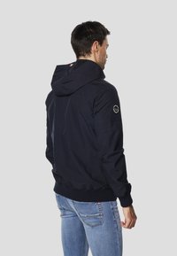 Lekka kurtka w kolorze granatowym z kapturem, elastycznymi mankietami i dolnym brzegiem. Posiada okrągłe logo na lewym ramieniu i gładką fakturę.