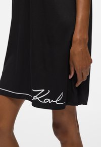 Rochie neagră din material moale, cu o linie accent curbată albă și logo-ul semnătură "Karl" la tiv. Lungime scurtă, croială lejeră.