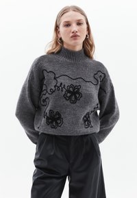 Maglione grigio lavorato a maglia con colletto alto, caratterizzato da ricami neri di fiori e viti. Abbinato a pantaloni neri larghi, con un focus su texture e design.