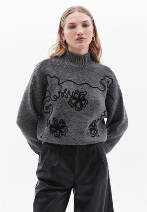 Grijze gebreide trui met hoge hals, versierd met zwarte bloemen- en wijnstokkenborduursels. Gestyled met losse zwarte broeken, met de nadruk op textuur en ontwerp.