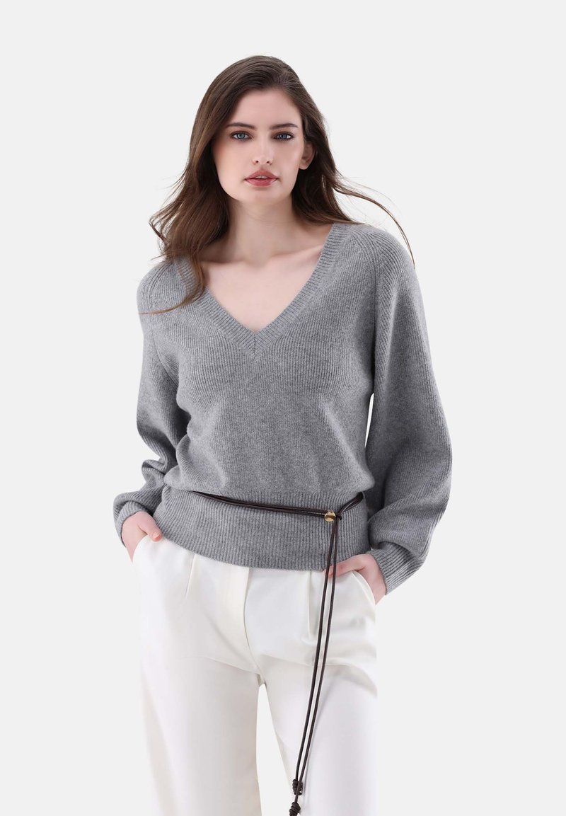 Grauer Strickpullover mit V-Ausschnitt und gerippter Textur. Lange Ärmel, lockere Passform, kombiniert mit weißen Hosen und einem braunen Kordelzuggürtel.