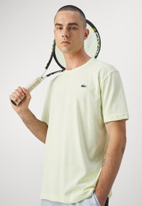 Lys grøn t-shirt i bomuld med korte ærmer, med et lille mørkegrønt logo på venstre bryst. Holder en tennisracket med gule detaljer.