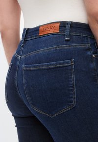 Blugi din denim albastru închis, cu talie înaltă, având o etichetă din piele cu inscripția „ONLY” cusută, și buzunare din spate contourate cu cusături bej.