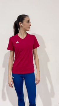 Femme portant un t-shirt rouge Adidas et un legging bleu, debout de profil et regardant vers sa gauche, avec des écouteurs sans fil dans les oreilles.