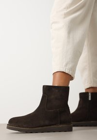 Botas de tobillo de ante marrón con punta redondeada y cremallera lateral, que cuentan con una superficie texturizada y una suela de goma baja y plana.
